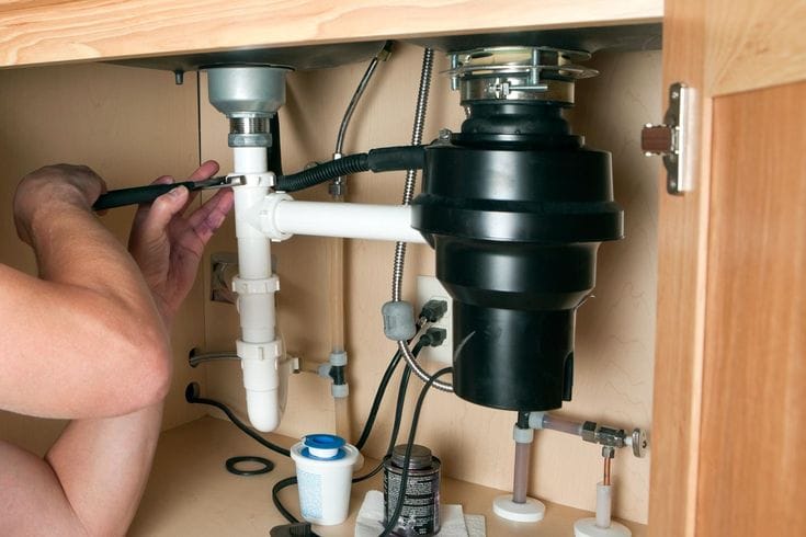 Plumber installing a garbage disposal beneath a kitchen sink in Cheltenham
